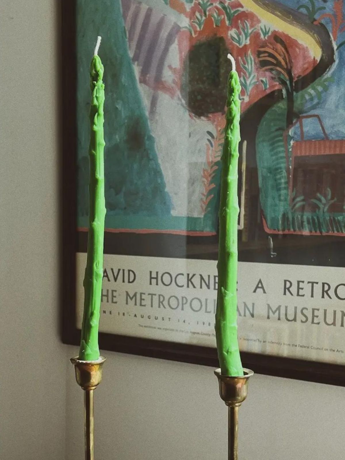 Two green candles in gold holders against a wall with a David Hockney poster.