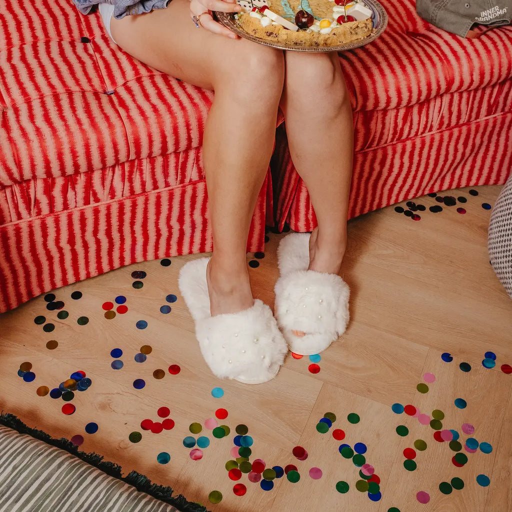 Person wearing white pearl fluffy slippers sitting on a red couch with colorful confetti on the floor.
